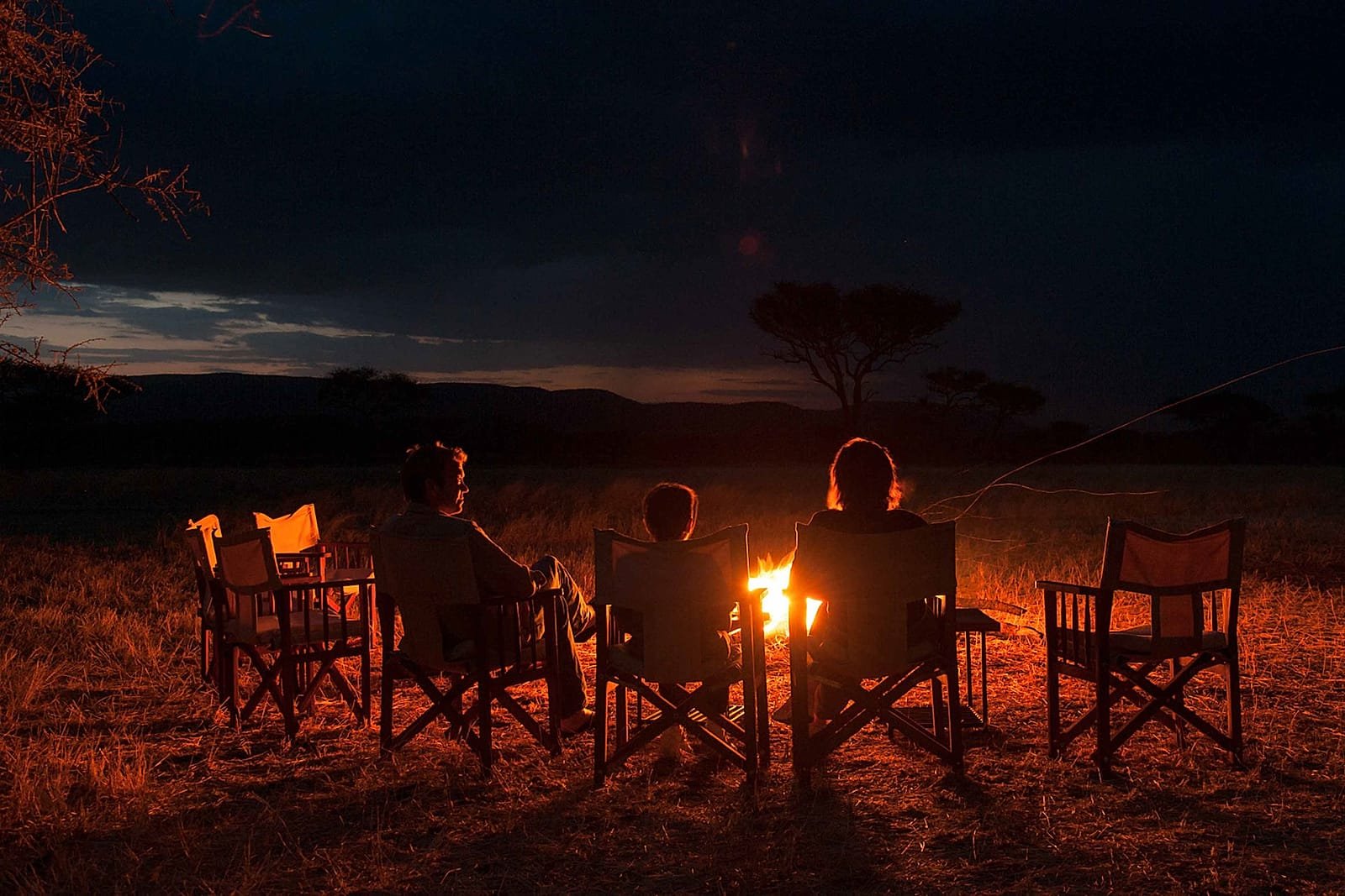 Guests in camp chairs around a fire after dark at a tented camp