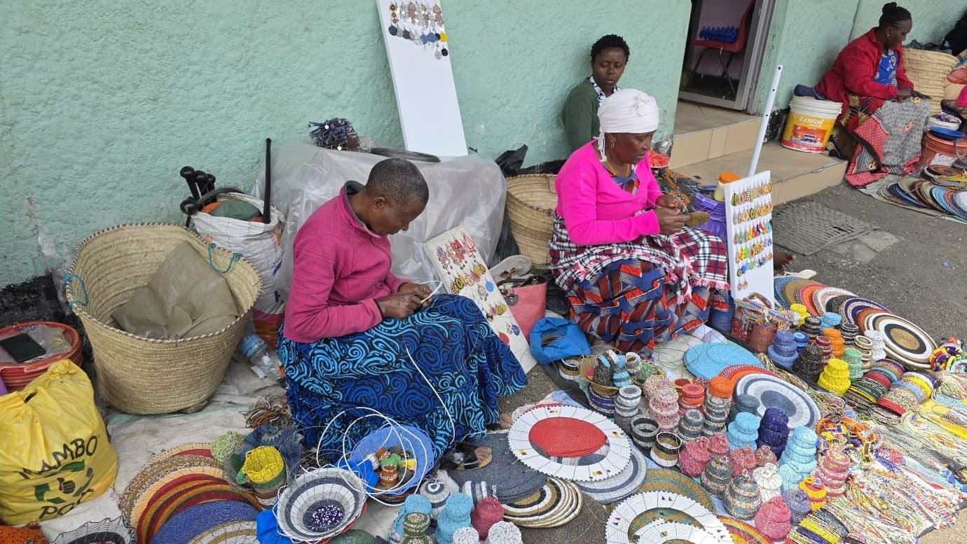 Two women stringing beadwork on the ground outside their shop, their bowls and coasters laid out around them